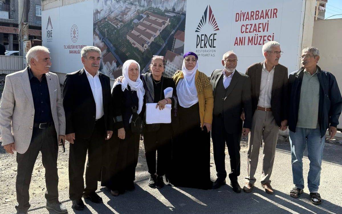 Fotoğrafta, sekiz kişiden oluşan bir grup, “Diyarbakır Cezaevi Anı Müzesi” yazısının bulunduğu bir inşaat alanı önünde yan yana duruyor. Arka planda Kültür ve Turizm Bakanlığı ile restorasyon çalışmasını yürüten Perge Restorasyonun logosu ve cezaevinin maket görüntüsü yer alıyor.