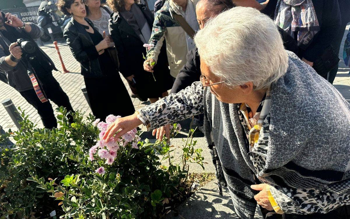 Fotoğrafta, güneşli bir günde bir grup insanın dış mekânda toplandığı görülüyor. Ön planda, yaşlı bir kadın elinde pembe çiçekler tutuyor ve bu çiçekleri yere, bir çalı bitkisinin üzerine bırakmak üzere eğiliyor.
