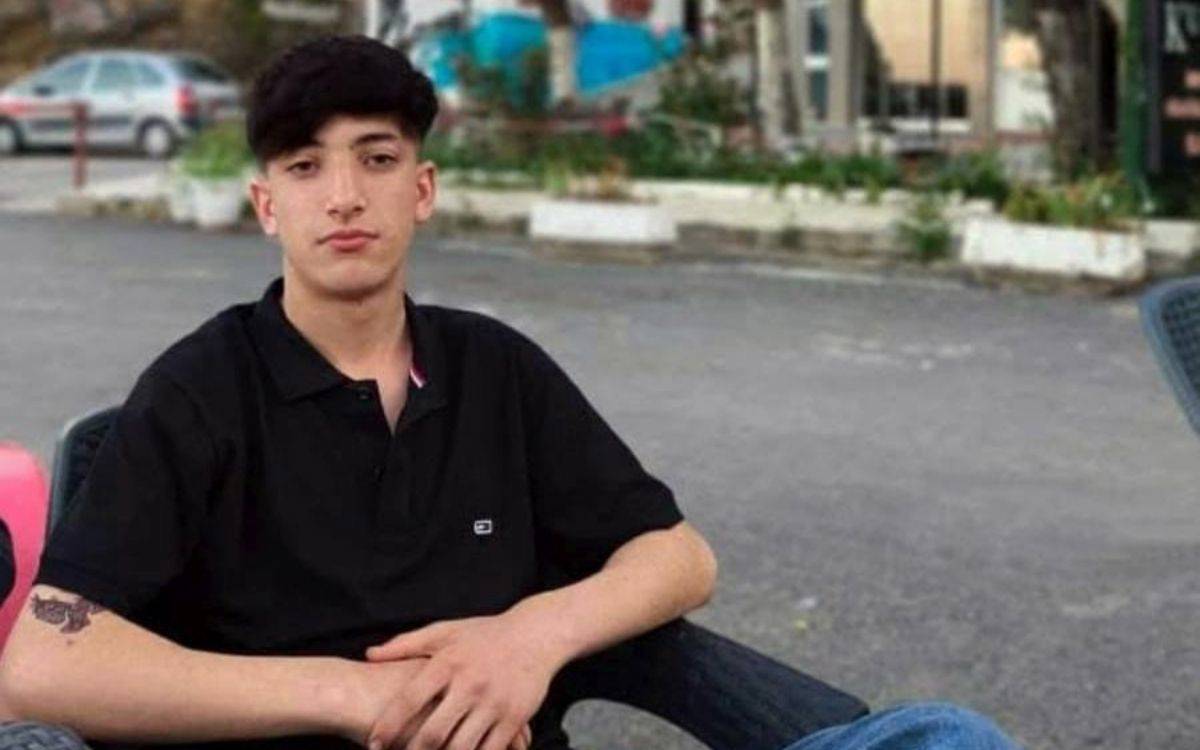 Fotoğrafta, açık havada bir sandalyede oturan genç bir erkek görülüyor. Kısa, koyu renkli saçları var ve siyah bir tişört giymiş. Kollarını rahat bir şekilde önünde birleştirmiş. Arka planda, yol kenarında park edilmiş bir araç, bitkiler ve bir bina dikkat çekiyor. Genel olarak fotoğraf, sakin ve gündelik bir atmosfer taşıyor.