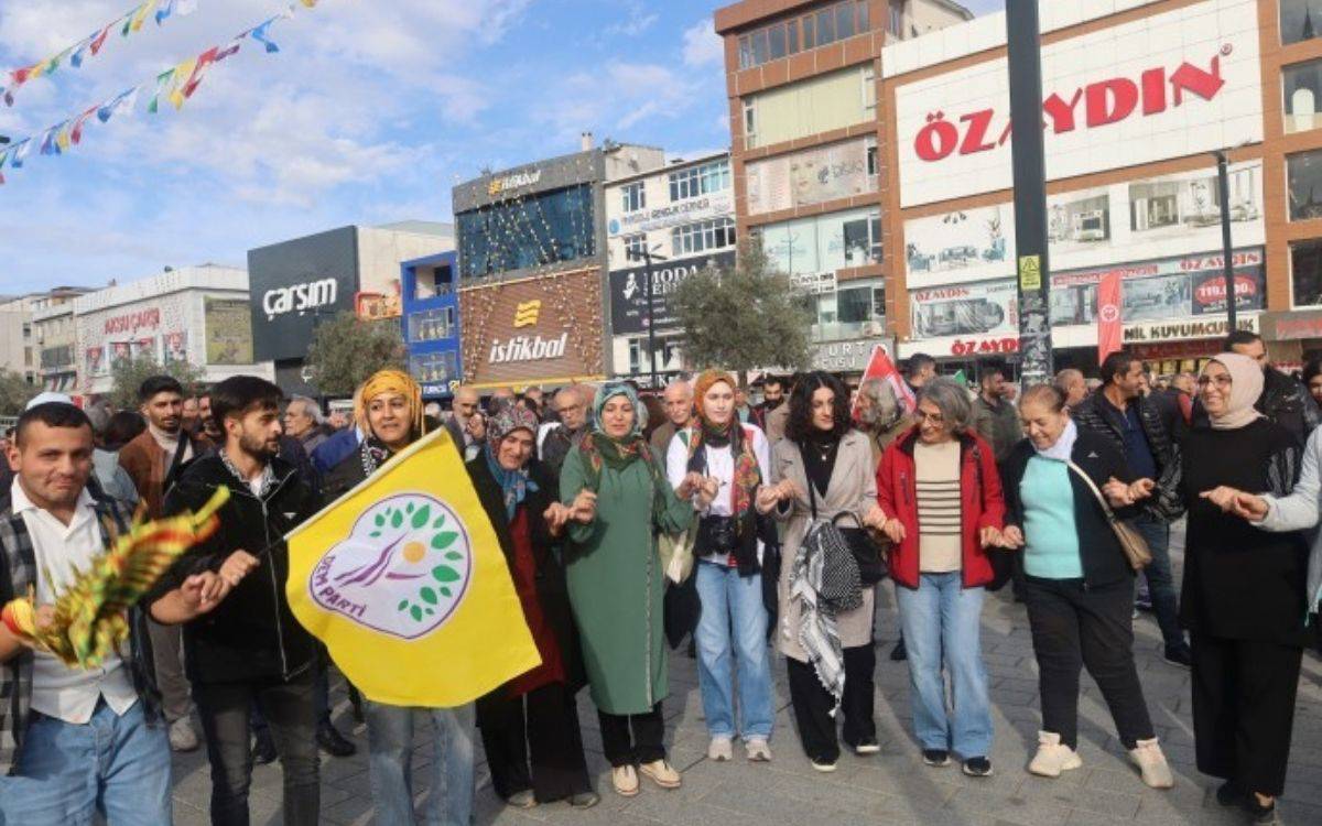 Fotoğrafta, açık bir alanda — muhtemelen bir meydanda — kalabalık bir grup insan el ele tutuşarak halay çekiyor. Ön sırada çoğunluğu kadınlardan oluşan bir grup yer alıyor; bazıları renkli başörtüleri takmış, bazıları başı açık. Orta kısımdaki bir kişi sarı zeminli, üzerinde yeşil yaprak ve mor yazı bulunan DEM Parti bayrağını tutuyor. Bayrağın üzerinde parti logosu net biçimde görülüyor.