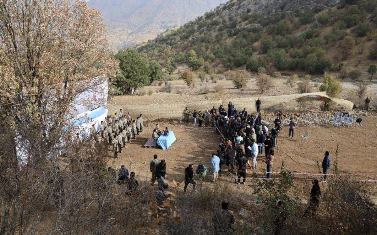 Fotoğrafta dağlık ve kayalık bir arazide, askeri üniforma giymiş bir grup insan yürürken görülüyor. Grup, arka plandaki taş merdivenlerden sırayla inerek dar bir patika üzerinde ilerliyor. Öndeki birkaç kişi kadın, diğerleri de karışık şekilde kadın ve erkeklerden oluşuyor.