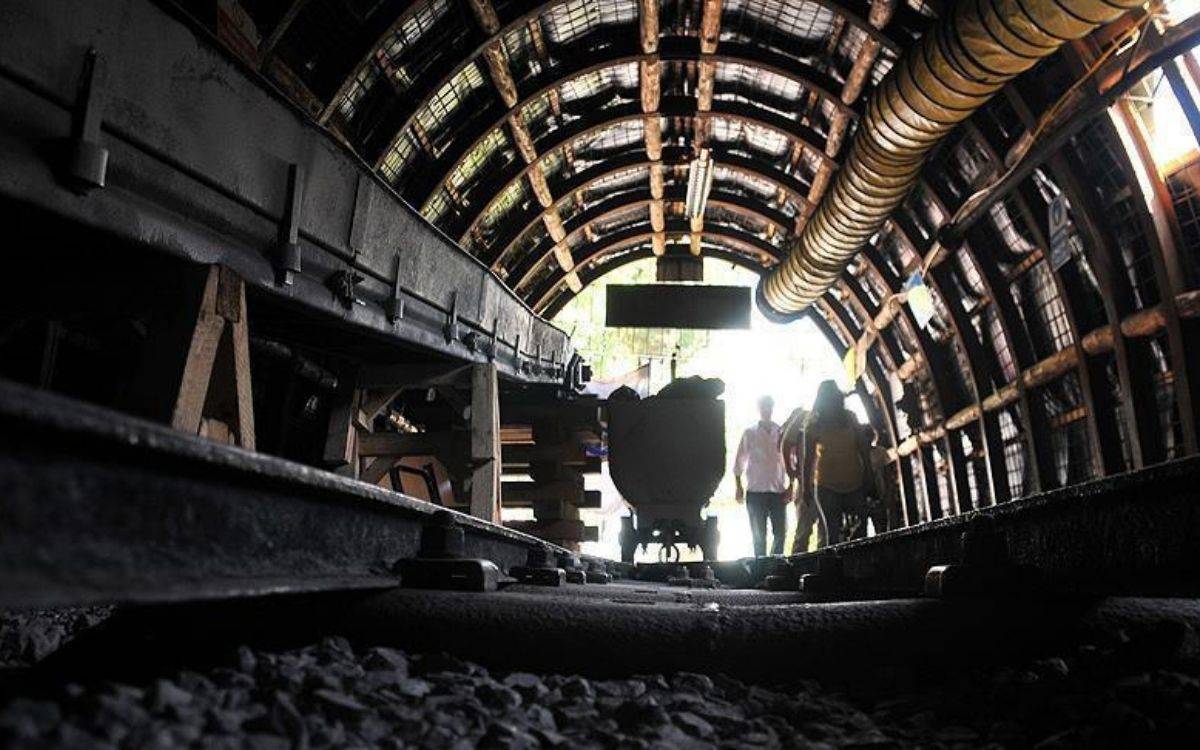 Fotoğrafta bir maden tünelinin içi görülüyor. Tünelin duvarları ve tavanı metal ve ahşap desteklerle güçlendirilmiş. Zeminde ray hattı uzanıyor ve raylar üzerinde maden vagonu bulunuyor. Tünelin sonunda, dışarıdan gelen gün ışığı görülüyor; bu kısımdan birkaç kişi içeri doğru yürümekte. Tünel içinde havalandırma boruları ve kablolar da dikkat çekiyor. Genel atmosfer, yer altındaki çalışma alanlarına özgü karanlık ve endüstriyel bir ortam yansıtıyor.