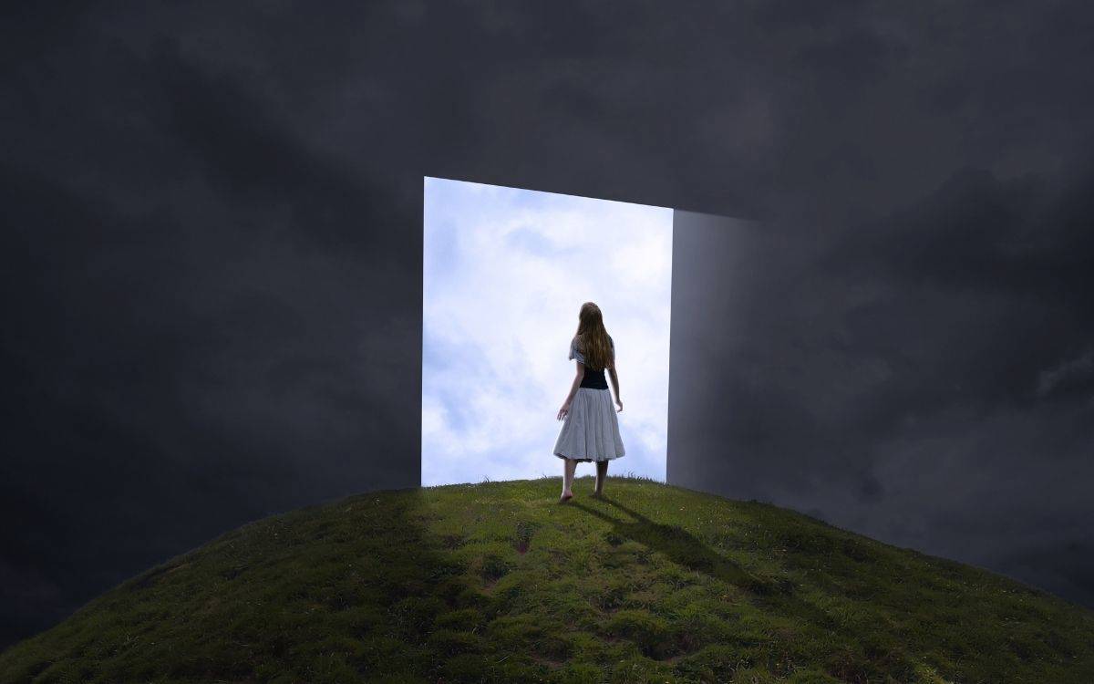 Fotoğrafta karanlık, yoğun bulutlarla kaplı bir gökyüzü altında yeşil bir tepe görülüyor. Tepede, sırtı kameraya dönük genç bir kadın duruyor. Üzerinde açık renkli bir etek ve koyu bir üst var, uzun saçları serbestçe sırtına dökülüyor.