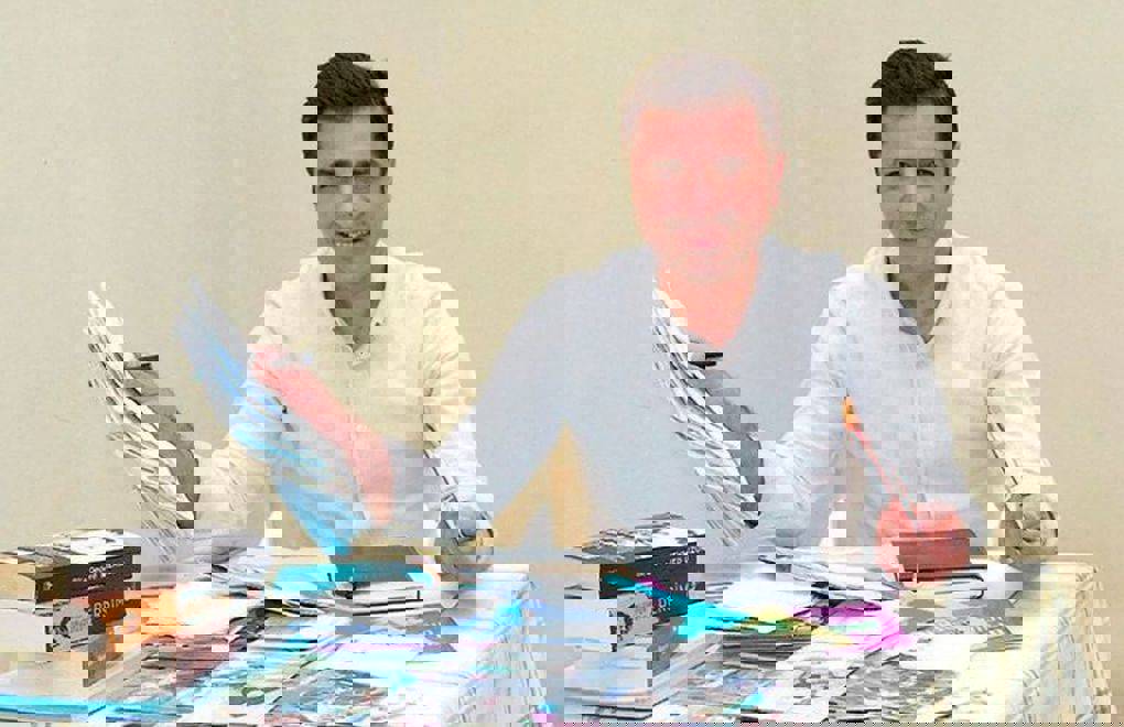 Fotoğrafta bir erkek, sade bir odada bir masa başında oturuyor. Üzerinde açık renkli, uzun kollu bir gömlek var. Gülümseyerek kameraya bakıyor. Masanın üzerinde kitaplar, defterler, mektuplar ve çeşitli kâğıtlar bulunuyor; kişi bu belgelerle ilgileniyor gibi görünüyor. Bu kişi Selahattin Demirtaş