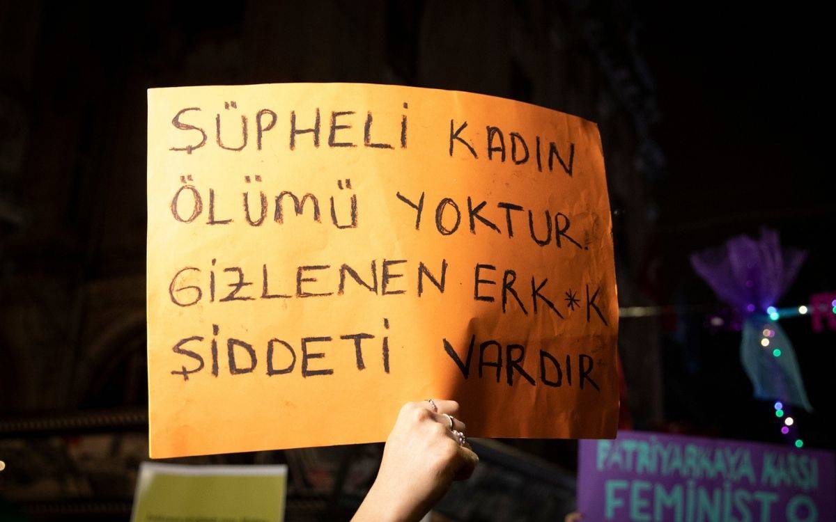 Fotoğrafta, gece saatlerinde düzenlenen bir protesto ya da yürüyüş sırasında çekilmiş bir sahne görülüyor. Önde, turuncu bir karton üzerine elle yazılmış bir pankart dikkat çekiyor. Pankartta siyah kalemle şu ifade yazıyor: “ŞÜPHELİ KADIN ÖLÜMÜ YOKTUR, GİZLENEN ERK*K ŞİDDETİ VARDIR.”