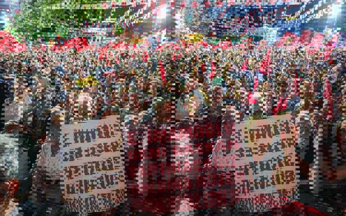 Kalabalık bir miting alanında, gece ışıkları altında sıkışık halde duran yüzlerce kişi görülüyor. Çoğunun elinde kırmızı bayraklar var; alanda flamalar ve süslemeler asılı. Ön sırada bariyerlere yaslanan bir grup dikkat çekiyor. İçlerinden biri büyük, beyaz zeminli bir pankart tutuyor: