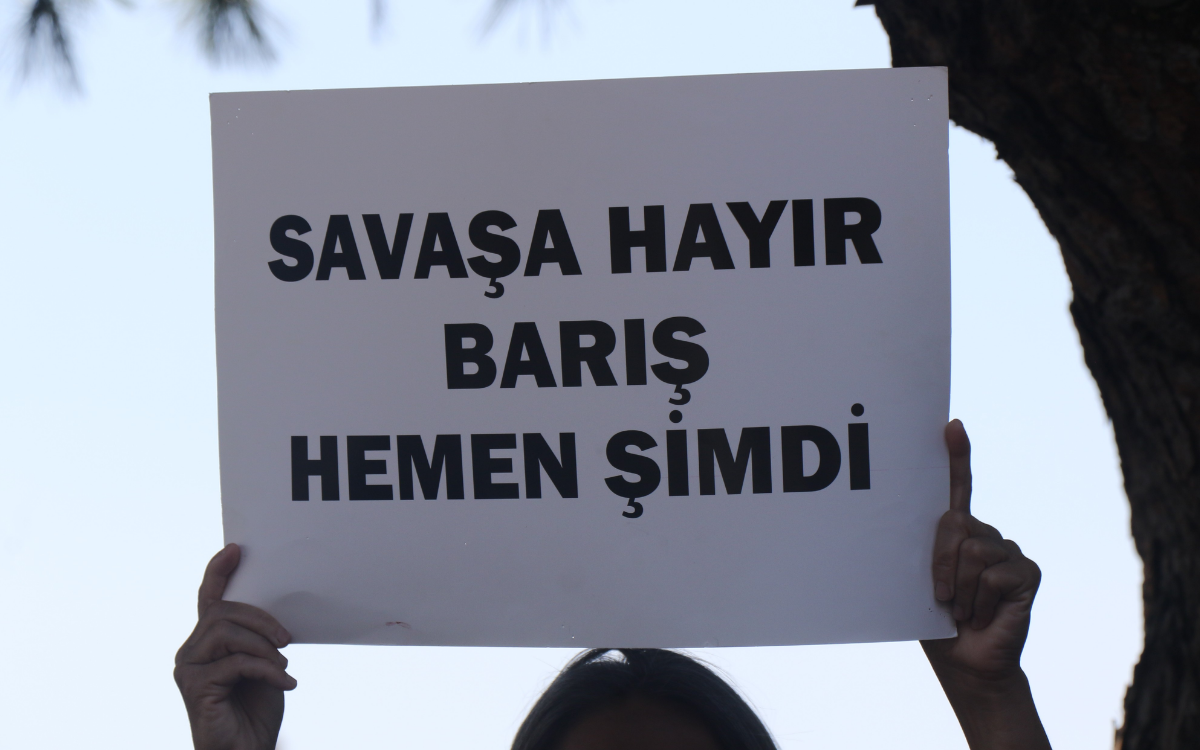 Beyaz bir kartona büyük siyah harflerle yazılmış bir pankart tutuluyor. Üzerinde üç satır halinde “SAVAŞA HAYIR / BARIŞ / HEMEN ŞİMDİ” yazıyor. Pankartı iki el yukarıdan kavramış; kişinin yüzü görünmüyor, sadece elleri ve başının üst kısmı kadraja giriyor. Arka planda ağaç gövdesi ve açık gökyüzü seçiliyor. Protesto/barış çağrısı temalı bir görüntü.