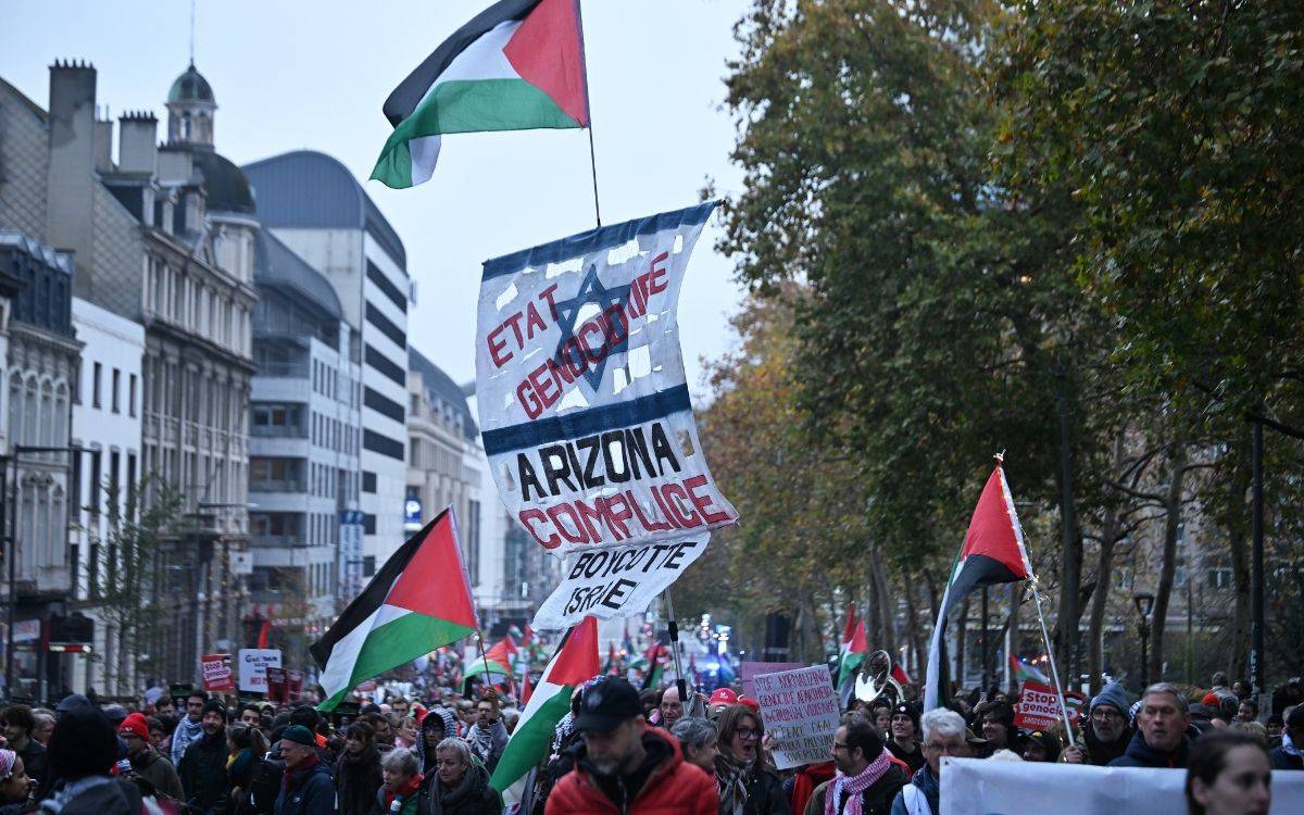 Fotoğrafta kalabalık bir protesto yürüyüşü görülüyor. Şehir merkezindeki geniş bir caddede toplanan göstericiler, ellerinde çok sayıda Filistin bayrağı taşıyor. Ön planda ve ortada büyük bir pankart dikkat çekiyor; üzerinde farklı dillerde yazılmış mesajlar ve ortasında bir sembol bulunuyor. Mesajlarda, İsrail’e yönelik eleştiriler ve boykot çağrıları yer alıyor.