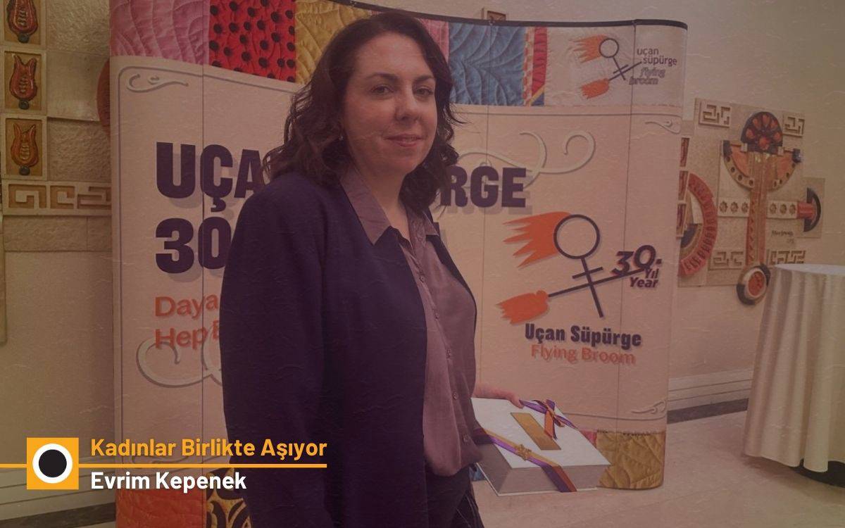 Fotoğrafta, kapalı bir etkinlik alanında bulunan yetişkin bir kadın gülümsüyor. Omuz hizasında koyu kahverengi dalgalı saçları var. Üzerinde mor tonlarında bir gömlek ve daha koyu mor bir ceket bulunuyor. Elinde renkli bir kurdeleyle bağlanmış bir dosya ya da belge tutuyor. Arka planda Uçan Süpürge / Flying Broom logosunun ve “30. Yıl” ibaresinin yer aldığı büyük bir pano görülüyor.