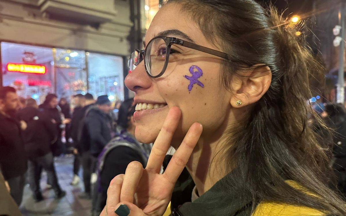 Fotoğrafta kalabalık bir sokakta, akşam saatlerinde çekilmiş neşeli bir an görülüyor. Ön planda genç bir kadın yer alıyor. Yüzünde büyük bir gülümseme var ve kameraya doğru barış işareti yapıyor. Kadının yanağında mor renkle çizilmiş bir kadın sembolü (♀) bulunuyor; bu, çoğunlukla kadın mücadelesi, eşitlik ve şiddete karşı dayanışmayı simgeleyen bir işaret. Gözlük takıyor, saçları arkaya doğru toplanmış, ışık sokak lambalarından ve etraftaki dükkânlardan geliyor.