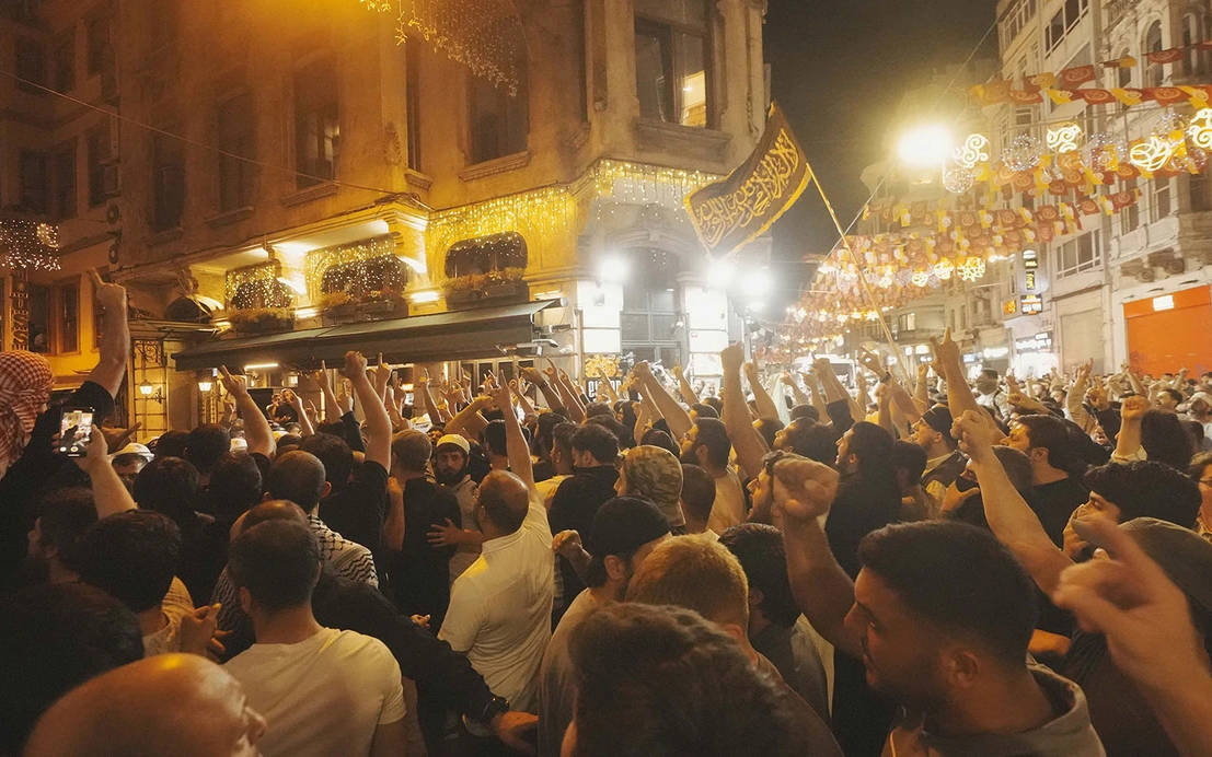 Gece saatlerinde çekilmiş bir sokak kalabalığı görülüyor. Dar bir cadde boyunca yüzlerce kişi bir araya gelmiş durumda. İnsanlar kollarını havaya kaldırmış, yumruklarını sıkmış ya da telefonlarıyla görüntü kaydediyor. Kalabalığın çoğunluğu erkeklerden oluşuyor.