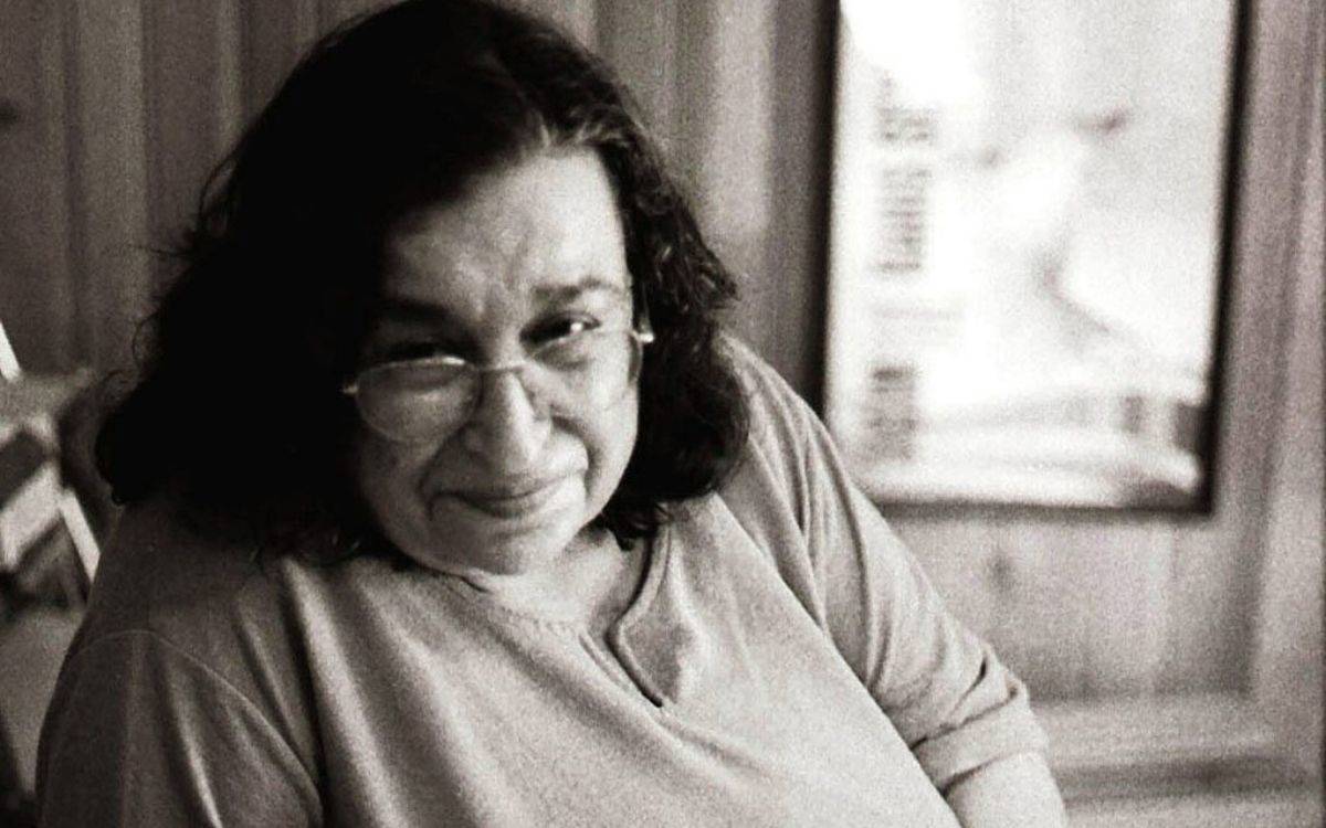 Fotoğraf siyah-beyaz tonlarda, sıcak ve samimi bir atmosferle çekilmiş. Kadrajın merkezinde, ileri yaşlarda bir kadın yer alıyor. Yüzündeki hafif gülümseme ve bakışlarının yumuşaklığı izleyene hemen bir yakınlık duygusu veriyor. İnce çerçeveli gözlükleri yüzüne rahatça yerleşmiş; saçları omuz hizasında, doğal hâliyle dalgalı.  Üzerinde sade, rahat bir bluz bulunuyor. Arka planda ahşap bir duvar ya da panel detayları seçiliyor, ayrıca flu biçimde bir afiş veya çerçeve görünmekte. Fotoğrafın genel havası hem içten, hem de derin bir duygu taşıyor; sanki yılların deneyimini, emeğini ve mücadelesini sessizce anlatan bir anın yakalanmış hâli.