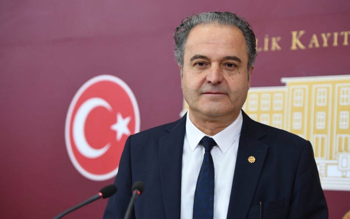 Görüntüde, resmi bir ortamda konuşma yapmak üzere mikrofonların önünde duran bir erkek görülüyor. Kişi takım elbise ve kravat giymiş, arka planda bordo renkte bir duvar ile üzerinde Türkiye Büyük Millet Meclisi’ni çağrıştıran bir amblem ve Türk bayrağı simgesi bulunuyor. Duvarın üzerinde altın renkli yazılar ve bir bina illüstrasyonu yer alıyor. Erkek, doğrudan kameraya bakarken sakin ve ciddi bir ifadeye sahip.