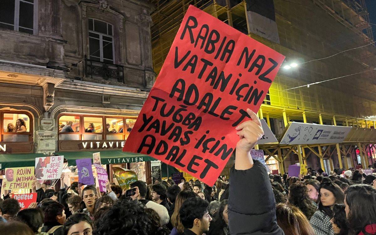 Fotoğrafta, kalabalık bir protesto veya yürüyüş sırasında insanların taşıdığı dövizler ve pankartlar görülüyor. Ön planda, bir kişinin elinde tuttuğu büyük, parlak kırmızı renkte bir pankart dikkat çekiyor. Bu pankartta büyük siyah harflerle adalet talep eden ifadeler yazılı. Pankart, kalabalığın üzerinde yükseltilmiş şekilde tutuluyor.