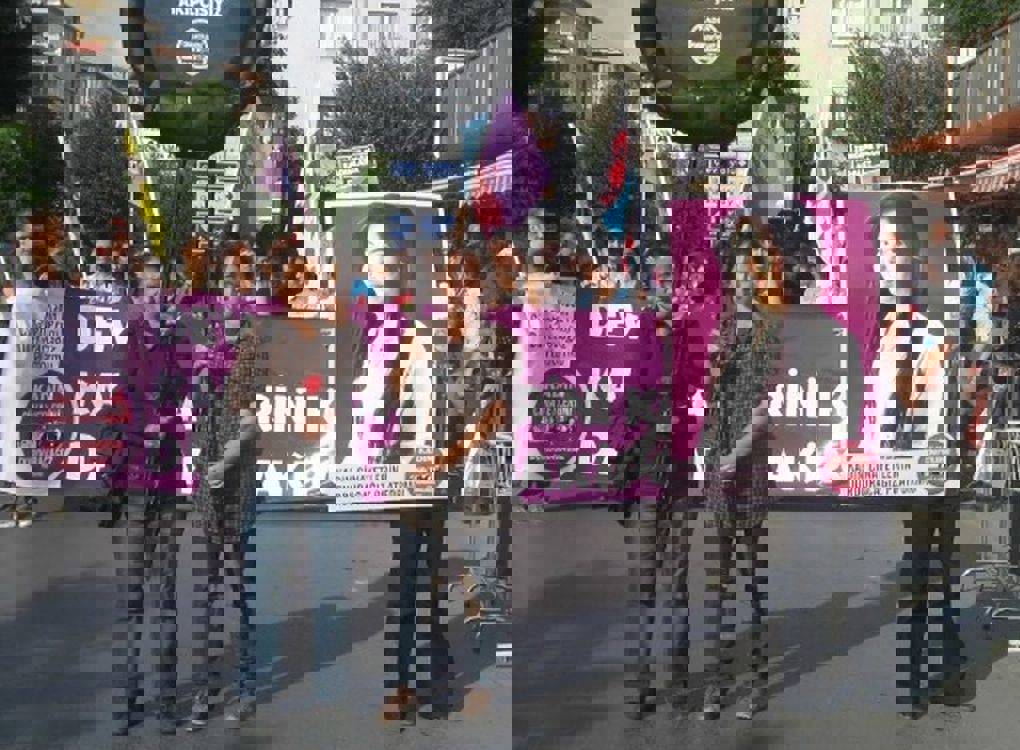 Görüntüde, sokakta gerçekleşen bir protesto yürüyüşü yer alıyor. Kalabalık bir grup, “Kadın Cinayetlerini Durduracağız Platformu” pankartları ve mor renkli büyük bir ana afiş taşıyor. Ön tarafta iki kadın öne çıkıyor: Biri elinde kırmızı bir karanfil tutuyor, diğeri büyük bir siyah çelenk taşıyor. Sağ tarafta ise başka bir kişi, genç bir kadının (fotoğrafta gülümseyen uzun saçlı bir kadın) büyük bir portresini taşıyor. Portrede ayrıca platformun logosu da bulunuyor.