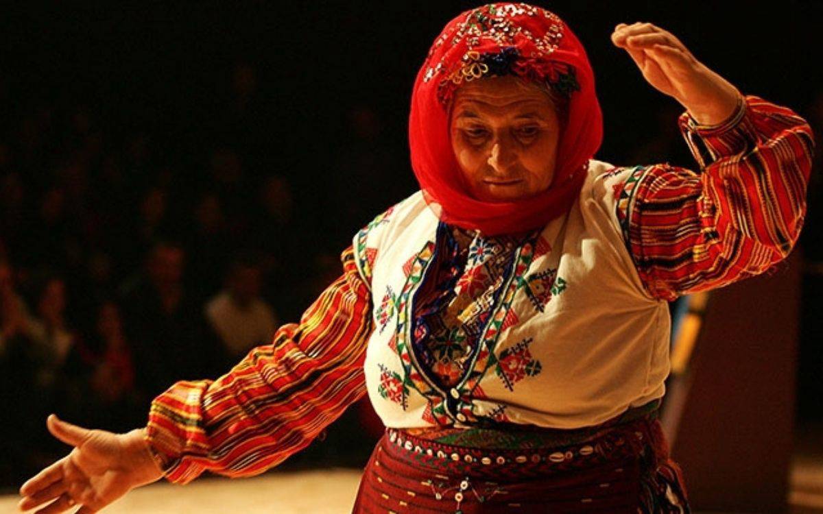 Görselde geleneksel kıyafetler giymiş yaşlı bir kadın sahnede dans ederken görülüyor.  Başında parlak kırmızı bir yazma/başörtüsü var; üzeri işlemeli, desenli ve ışıltılı detaylarla süslenmiş.