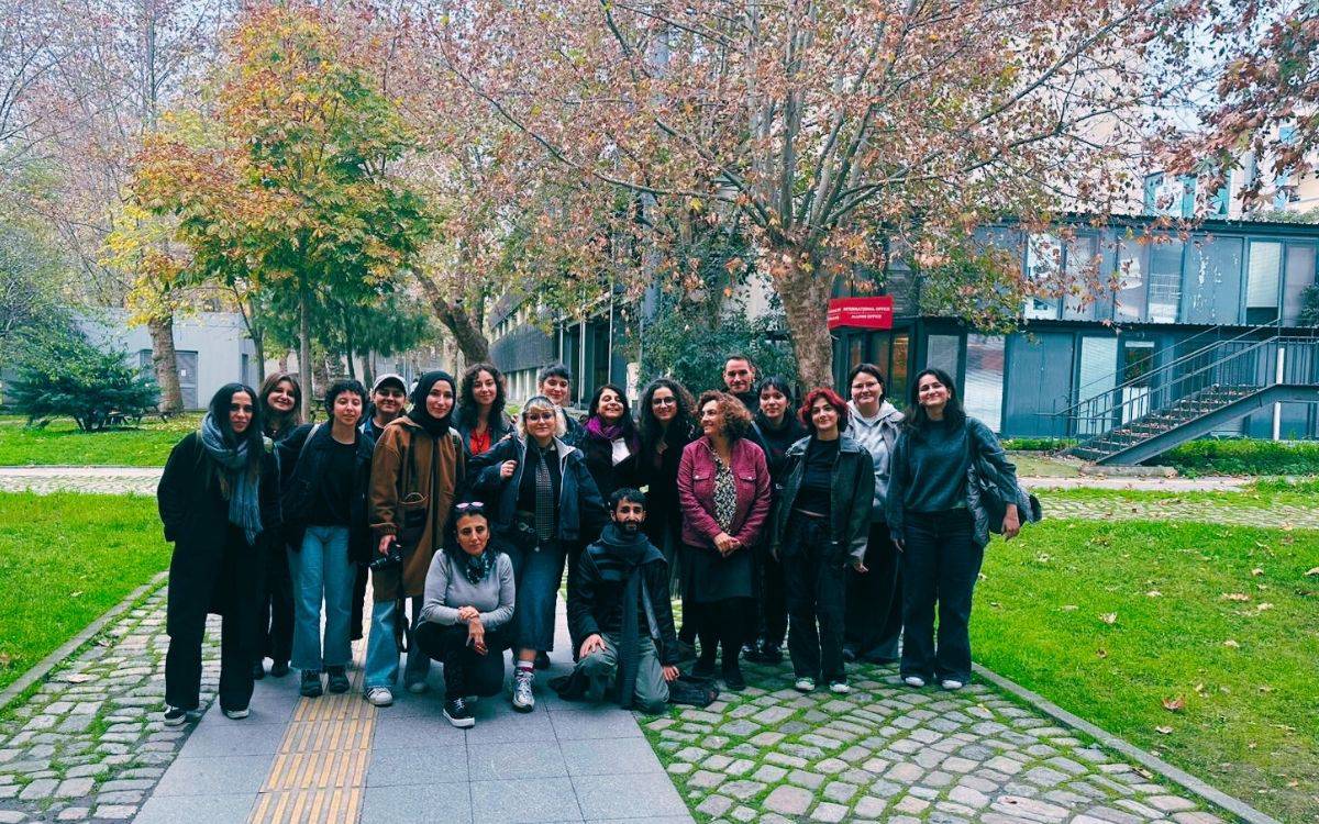 Görüntü, açık havada çekilmiş büyük bir grup fotoğrafı. Sonbahar yapraklarının döküldüğü, ağaçların sarı-kahverengi tonlara büründüğü yeşillikler içinde bir kampüs alanında çekilmiş. Arkada modern yapılar ve camlı bir bina ile dış merdiven görülüyor. Ön planda yaklaşık yirmi kişilik bir grup yan yana dizilmiş hâlde duruyor; iki kişi önde çömelmiş, diğerleri ayakta. Grupta farklı yaşlarda ve farklı tarzlarda giyinmiş insanlar var.