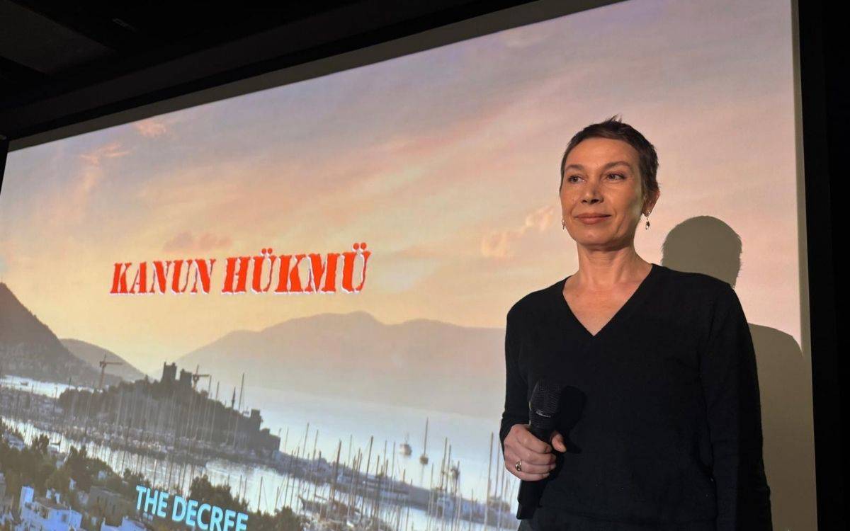 Fotoğrafta bir sinema salonu ya da etkinlik alanında çekilmiş bir sahne görülüyor. Ön planda, elinde mikrofon tutan bir kadın ayakta duruyor. Siyah, s