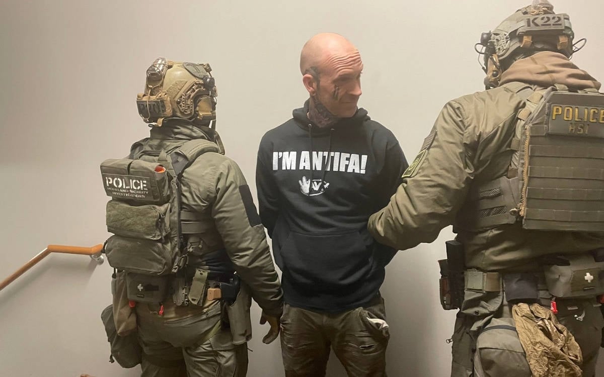 Bir odada, ortada elleri arkadan kelepçeli bir erkek duruyor. Siyah kapüşonlusunda “I’M ANTIFA!” yazıyor. Sağında ve solunda, yüzleri kasklı, ağır teçhizatlı iki polis onu tutuyor. Ortam sade, duvar açık renk.