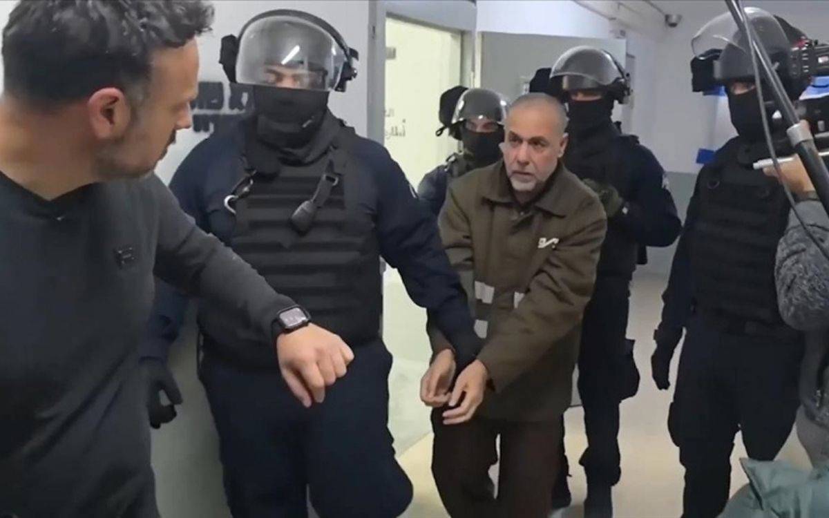 Fotoğrafta kapalı bir mekânda gerçekleşen bir gözaltı veya operasyon anı görülüyor. Ortada bulunan bir erkek kişinin elleri öne doğru uzatılmış ve muhtemelen kelepçeli durumda. Üzerinde kahverengi bir mont var ve yüzünde gergin bir ifade bulunuyor.