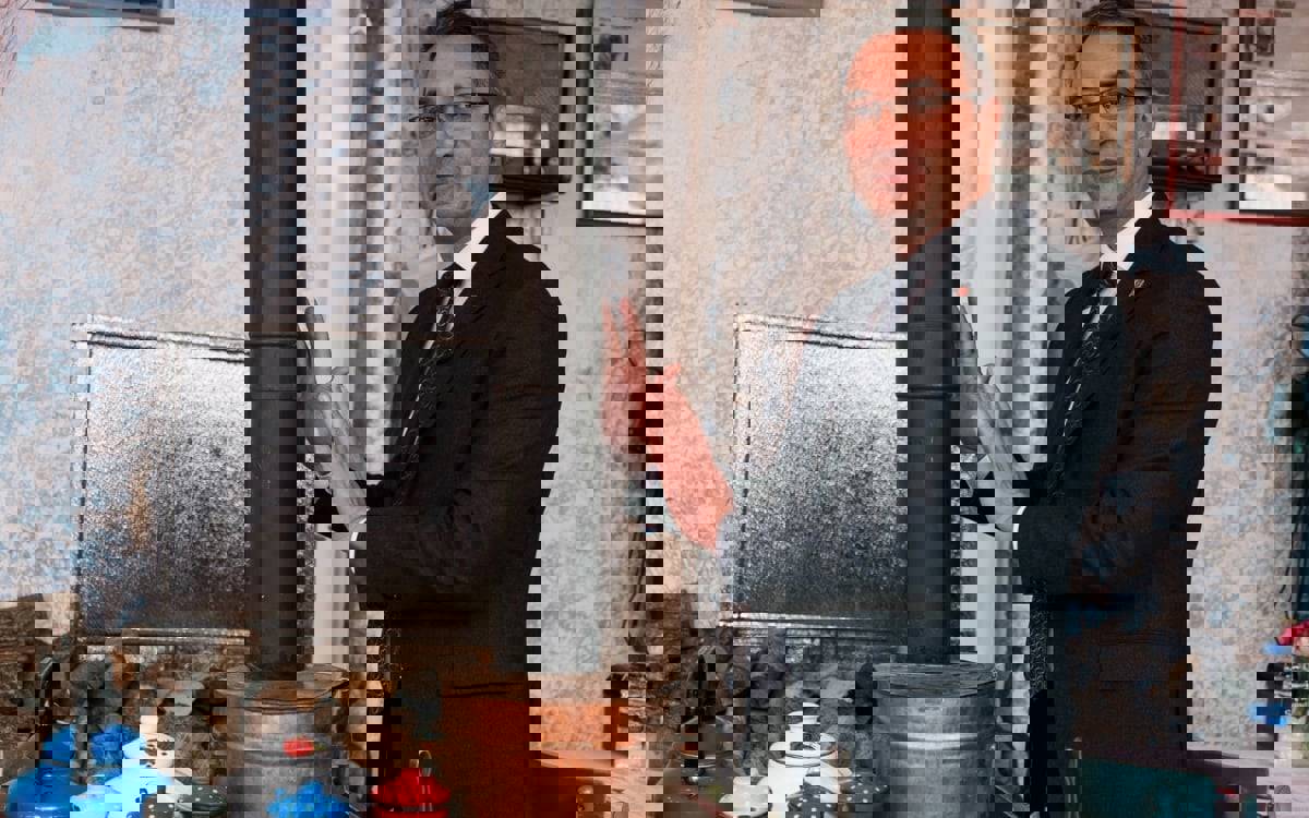 Fotoğrafta takım elbise, gömlek ve kravat giymiş orta yaşlı bir erkek iç mekânda ayakta duruyor. Hafifçe gülümseyerek kameraya bakıyor ve ellerini siyah borulu, bakır renkli gövdeli bir sobaya doğru uzatmış. Arka planda açık renkli, desenli bir duvar; duvara asılı eski tip bir radyo ya da ahşap kutu görünümlü cihaz; çerçeveli bir belge ya da gazete kupürü bulunuyor.