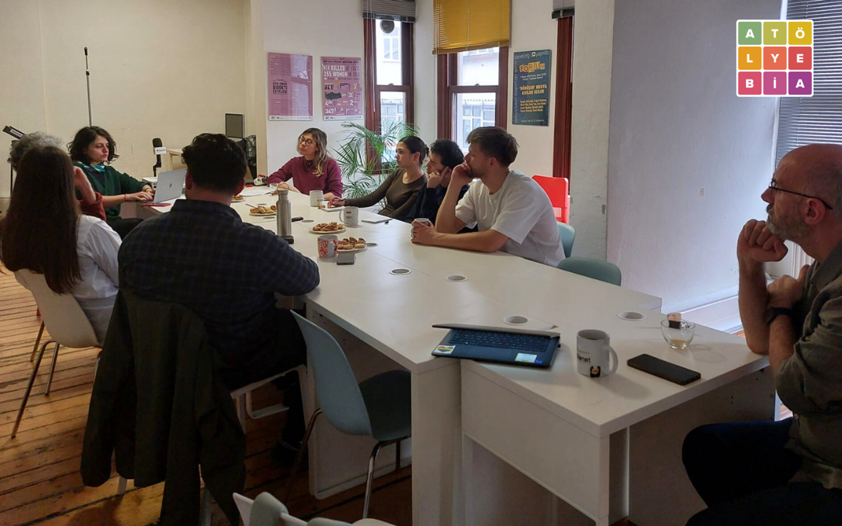 Bir toplantı odasında, uzun bir masa etrafında oturan bir grup insanın katıldığı küçük bir atölye/çalışma buluşması görülüyor. Katılımcılar konuşanı dikkatle dinlerken masada kahve kupaları, atıştırmalıklar ve dizüstü bilgisayarlar yer alıyor; ortam samimi ve odaklı bir çalışma havası taşıyor.