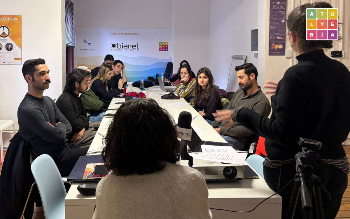 Bir toplantı odasında gerçekleştirilen gazetecilik atölyesi görülüyor. Uzun bir masanın etrafında oturan katılımcılar konuşmacıyı dikkatle dinlerken, sağ tarafta ayakta duran bir eğitmen anlatım yapıyor. Masada dizüstü bilgisayarlar, notlar ve bir projeksiyon cihazı bulunuyor; arkadaki duvarda bianet / IPS İletişim Vakfı logosunun yer aldığı bir pano görülüyor. Ortam, küçük bir eğitim veya atölye çalışması atmosferini yansıtıyor.