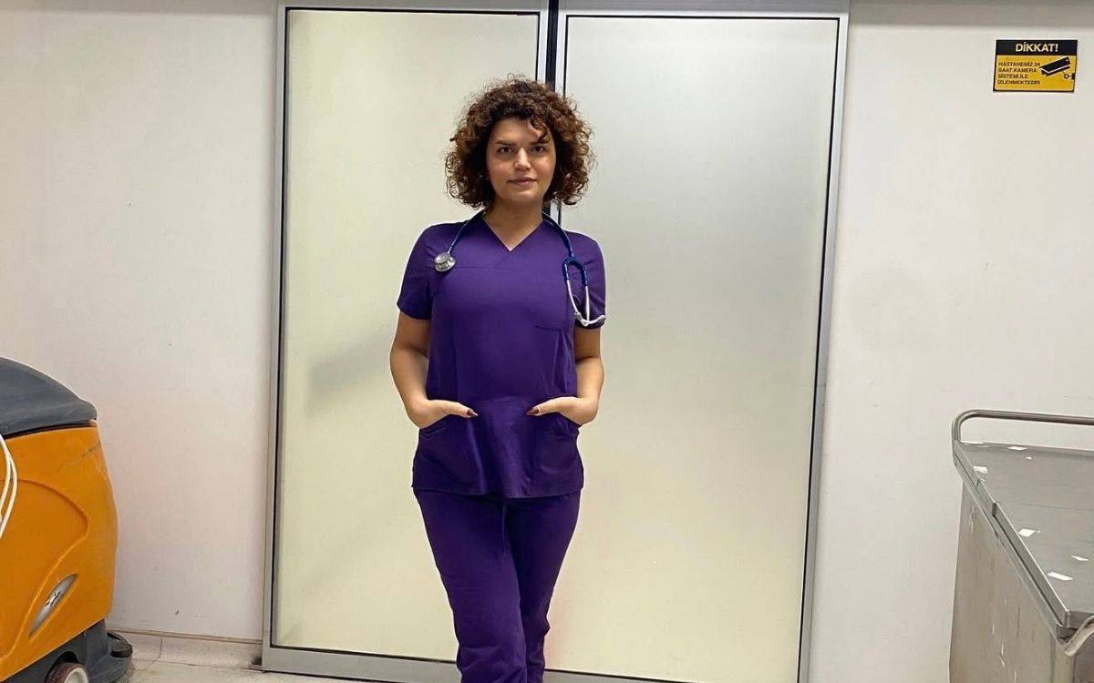 Mor scrubs giymiş, boynunda stetoskop olan kıvırcık saçlı bir kadın hastane koridorunda duruyor; elleri ceplerinde, kameraya sakin ve kendinden emin bakıyor.