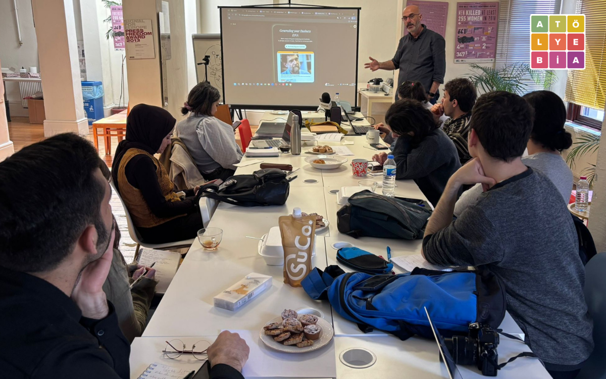 Bir toplantı masası etrafında oturan katılımcılar, projeksiyon ekranı önünde sunum yapan bir konuşmacıyı dinliyor. Masada bilgisayarlar, not defterleri, kameralar ve atıştırmalıklar bulunurken, arka planda Atölye BİA logosu ve gazetecilikle ilgili afişler yer alıyor.