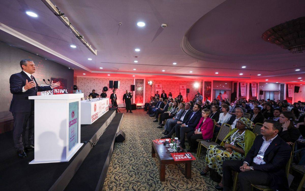 Bir konferans salonunda kürsüde konuşan bir siyasetçi, mikrofonların önünde elini kaldırarak hitap ediyor; karşısında oturan kalabalık bir dinleyici grubu dikkatle dinliyor, salonda kırmızı ışıklandırma hâkim.