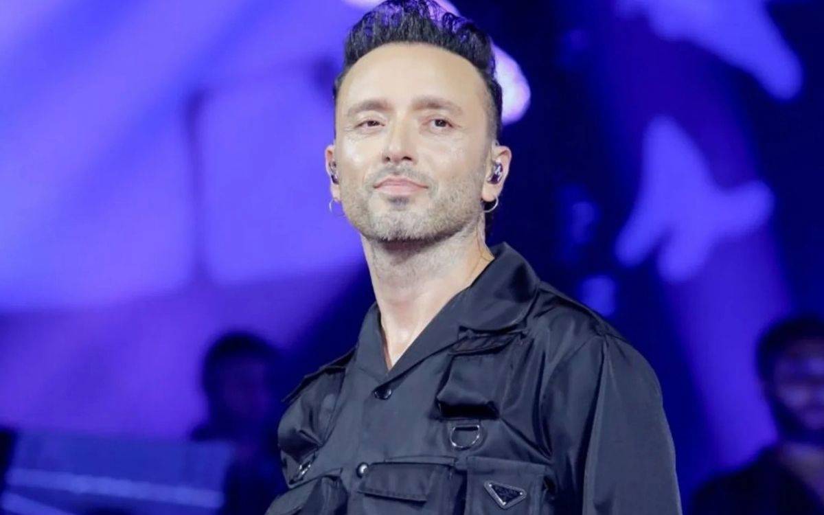 Sahne ışıkları altında siyah kıyafetli bir sanatçı, kulak içi kulaklıkla kameraya doğru bakıyor; arka planda mor ve mavi tonlarda konser ışıkları görülüyor.