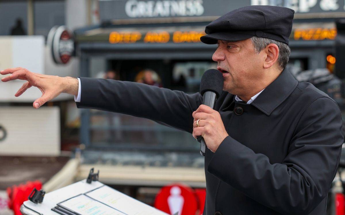 Şapkalı, koyu paltolu bir erkek açık havada mikrofona konuşurken kolunu ileri uzatıyor; önünde kürsü, arka planda dükkân tabelaları bulanık.