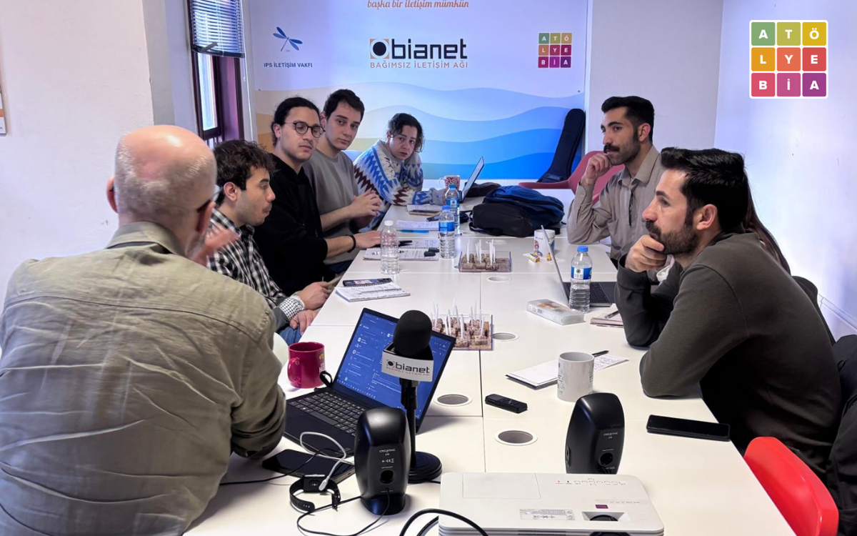 Bir toplantı masası etrafında toplanan atölye katılımcıları, bir konuşmacıyı dinlerken notlar alıyor. Arka planda IPS İletişim Vakfı, bianet ve Atölye