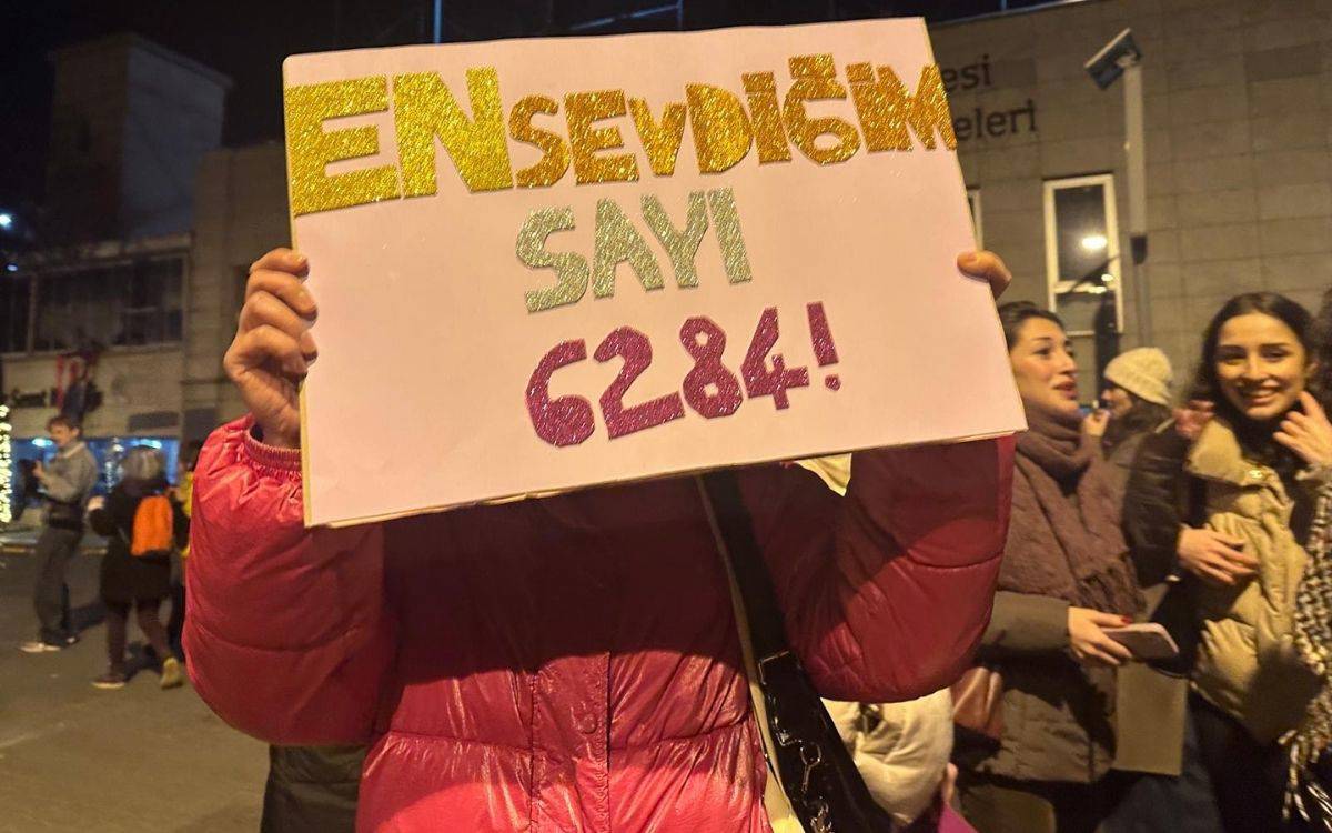 Gece sokakta pembe montlu bir kişi, yüzünü kapatan pankart tutuyor. Simli harflerle “EN sevdiğimiz sayı 6284!” yazıyor; arka planda kalabalık görünüyor.