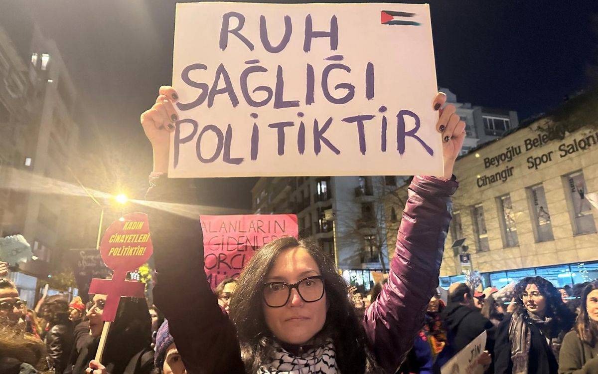 Geceki kalabalık eylemde gözlüklü bir kadın, başının üstünde “Ruh sağlığı politiktir” yazılı pankart tutuyor. Arkada mor feminist simgeler ve protestocular var.
