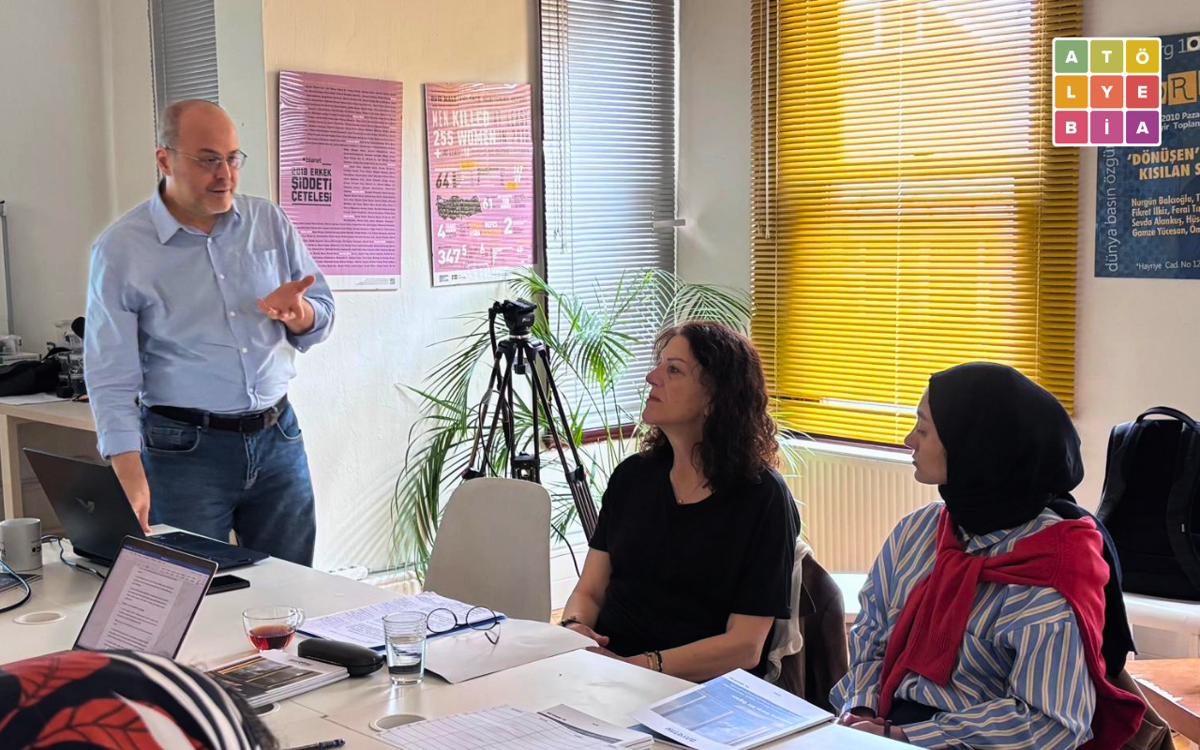 Atölye BİA’da düzenlenen etkinlikte ayakta konuşan bir erkek katılımcılara sunum yapıyor. Masada oturan iki kadın konuşmacıyı dikkatle dinlerken, masa