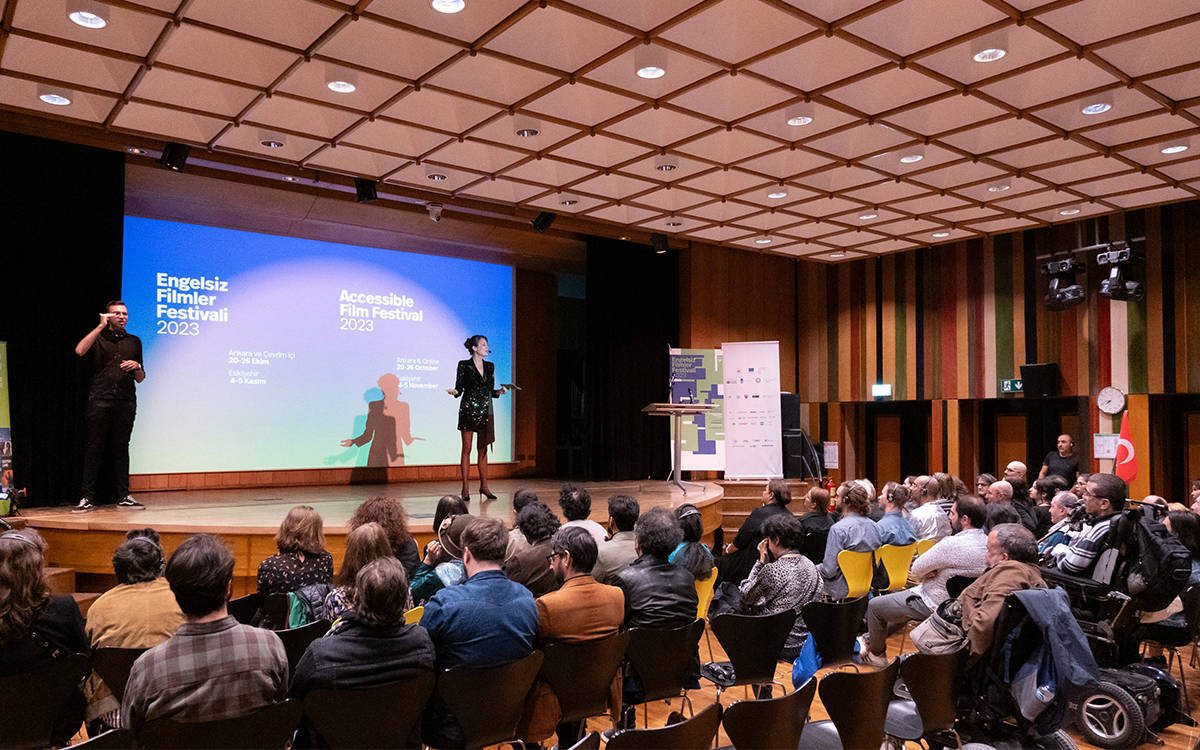 Sahnenin sol tarafında siyah giyimli bir işaret dili tercümanı ayakta duruyor ve çeviri yapıyor. Sahnenin ortasında ise elinde notlar tutan bir sunucu konuşma yapıyor. Sunucunun arkasındaki büyük ekranda mavi-beyaz degrade bir görsel üzerinde “Engelsiz Filmler Festivali 2023” ve “Accessible Film Festival 2023” yazıları yer alıyor. Ekranda ayrıca etkinliğin Ankara ve çevrim içi ayağı için 20–26 Ekim, Eskişehir ayağı için ise 4–5 Kasım tarihleri belirtilmiş.