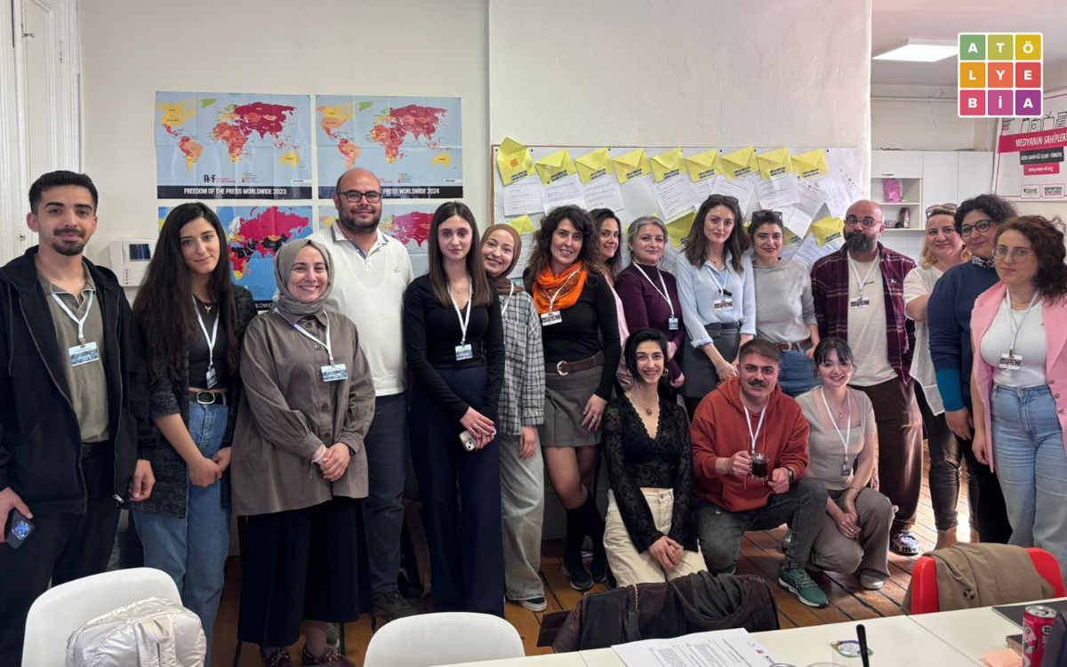 Bir atölye ortamında çekilmiş toplu fotoğraf görülüyor. Farklı yaş ve mesleklerden katılımcılar yan yana poz veriyor; çoğunun boynunda yaka kartı var. Arka duvarda basın özgürlüğü haritaları ve notların asılı olduğu bir pano yer alıyor. Sağ üstte Atölye BİA logosu bulunuyor.