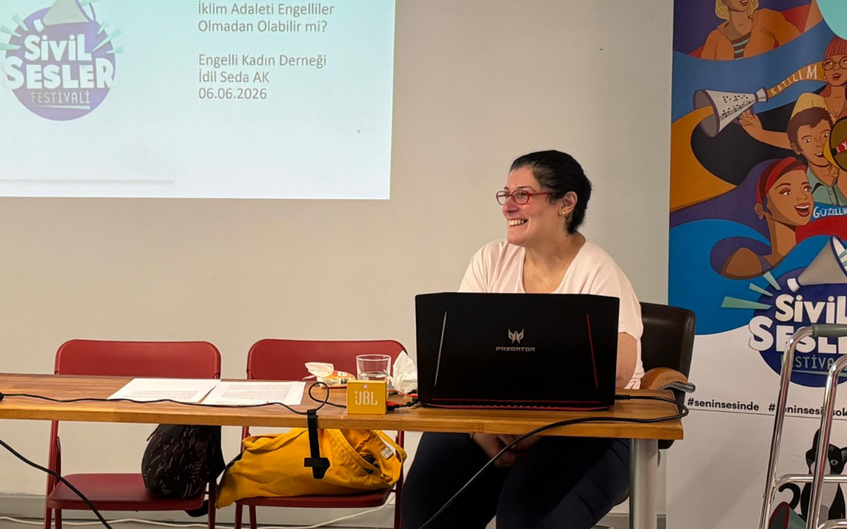 Görselde, Sivil Sesler Festivali kapsamında düzenlenen "İklim Adaleti Engelliler Olmadan Olabilir mi?" başlıklı atölyeyi yürüten İdil Seda Ak bir masanın arkasında otururken görülüyor. Önündeki dizüstü bilgisayar üzerinden sunum yapan Ak gülümserken, arkasındaki projeksiyon ekranında atölyenin başlığı ve tarih bilgisi yer alıyor. Sağ tarafta ise Sivil Sesler Festivali afişi görülüyor.