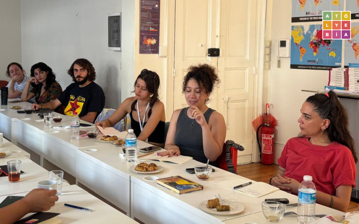 Katılımcılar, Atölye BİA’nın Beyoğlu’ndaki ofisinde masa etrafında otururken görülüyor. Masalarda not defterleri, içecekler ve yiyecekler bulunuyor.