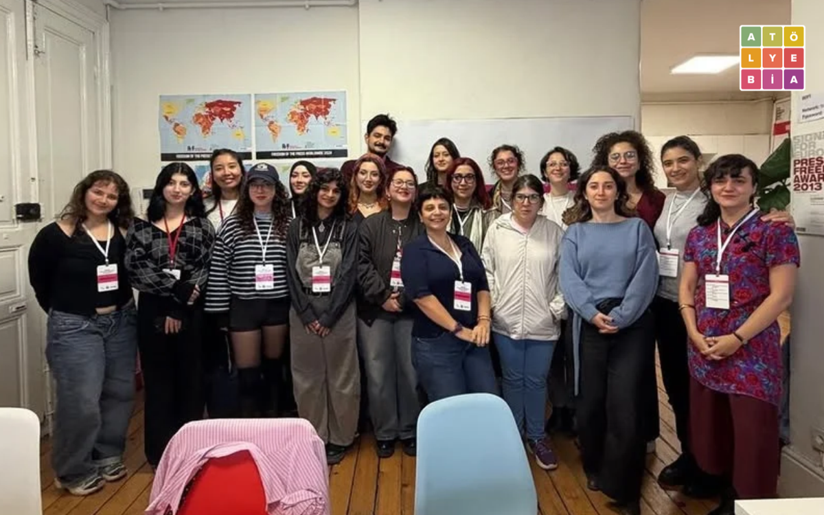 Bir atölye salonunda, yaklaşık 20 kişilik bir grup yan yana durarak kameraya poz veriyor. Arka planda dünya haritaları ve beyaz tahta, sağ üst köşede Atölye BİA logosu görülüyor.