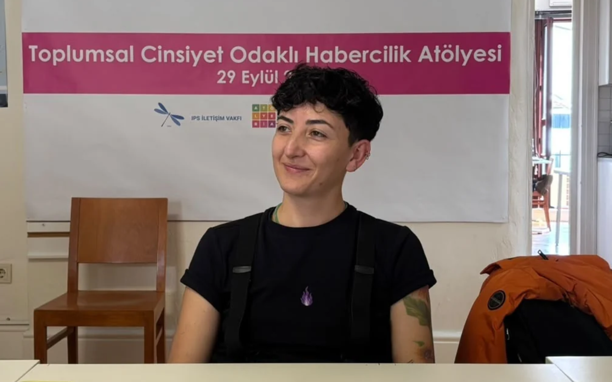 Kısa siyah saçlı, siyah tişörtlü ve kollarında dövmeler bulunan bir kişi, masada otururken gülümsüyor. Arkasında “Toplumsal Cinsiyet Odaklı Habercilik Atölyesi” yazılı Atölye BİA afişi yer alıyor.
