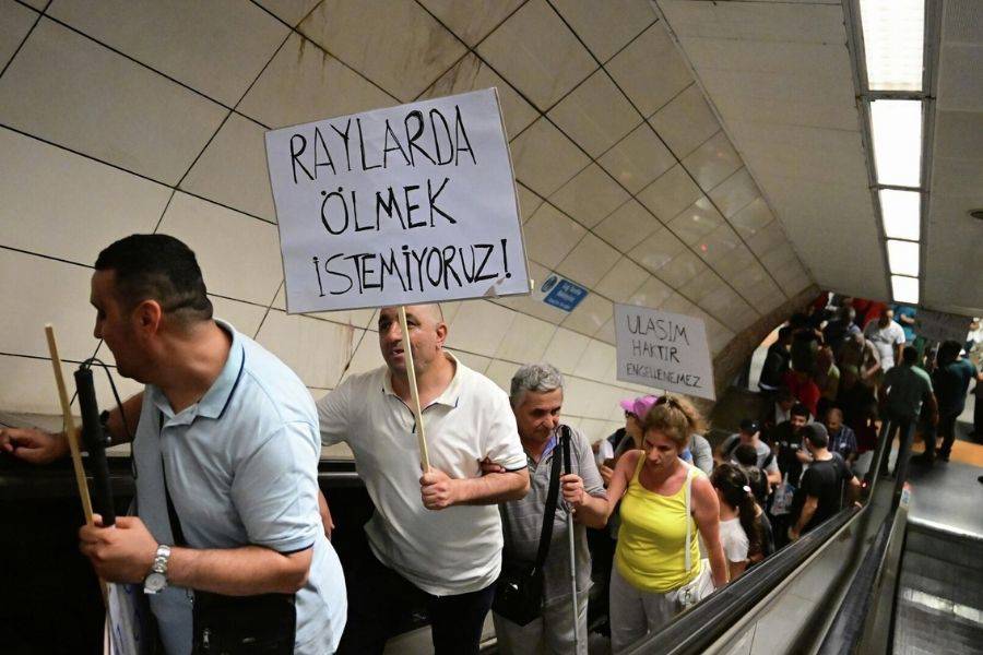 Fotoğrafta, bir metro istasyonunda düzenlenen bir protesto gösterisi görülüyor. Bir grup insan, yürüyen merdivenlerden yukarı doğru çıkarken ellerinde pankartlar taşıyorlar. En önde bulunan kişi, üzerinde "Raylarda ölmek istemiyoruz!" yazılı bir pankart tutuyor. Arkasından gelen bir başka kişinin taşıdığı pankartta ise "Ulaşım haktır, engellenemez" yazılı. Protestocular kararlı bir şekilde ilerliyor ve yüz ifadeleri ciddi. Fotoğraf, toplu taşıma güvenliği ve ulaşım hakkı ile ilgili bir talepleri dile getiren bir protesto anını yansıtıyor. Mekan, tipik bir metro istasyonu olup duvarlarda fayanslar ve aydınlatmalar görülüyor. Kalabalık, protesto esnasında topluca hareket ediy