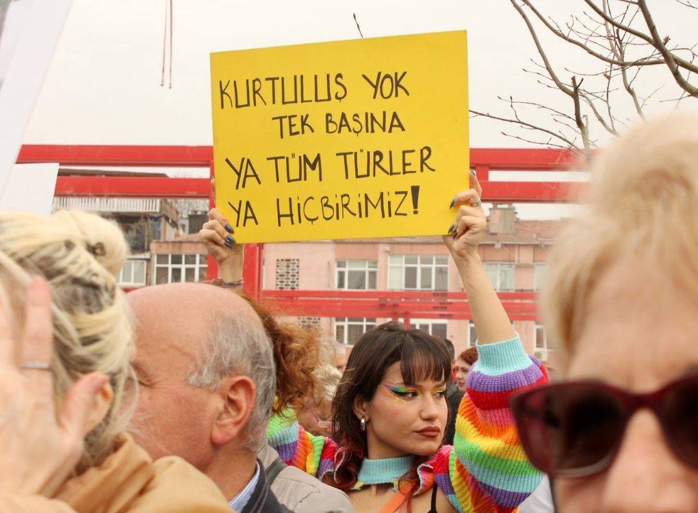 Görselde, bir protesto veya mitingde yer alan bir grup insan görülüyor. Önde duran genç bir kadın, elinde sarı bir pankart tutuyor. Pankartta, "Kurtuluş yok tek başına, ya tüm türler ya hiçbirimiz!" yazılı.