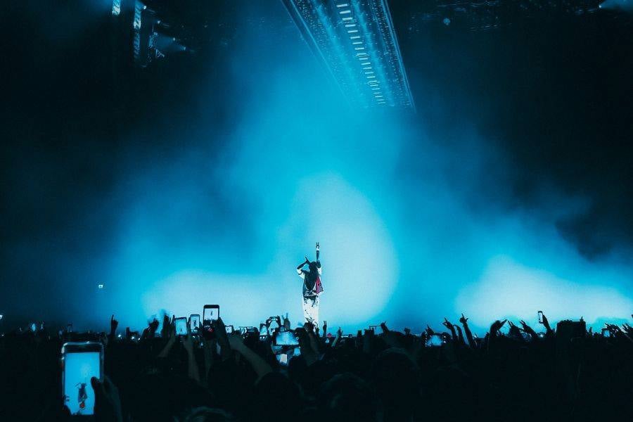 Fotoğrafta, büyük bir konser veya müzik etkinliğinde sahnede performans sergileyen bir sanatçı görülüyor. Sahne mavi bir sisle kaplanmış ve bu sis sahneye dramatik bir atmosfer katıyor. Sanatçı, sahnenin ortasında tek başına duruyor ve bir elini havaya kaldırmış, bu da güçlü bir sahne duruşu yaratıyor. Seyirciler, ellerindeki telefonlarla bu anı kaydetmeye çalışıyor ve kalabalığın arasında yükselen telefon ekranları dikkat çekiyor. Sahne ışıkları, sanatçının siluetini ön plana çıkarırken arka plandaki sis, sahne atmosferini yoğunlaştırıyor. Genel olarak, fotoğraf sahnenin enerjisini ve izleyicilerin coşkusunu yansıtan dinamik bir anı yakalamış.