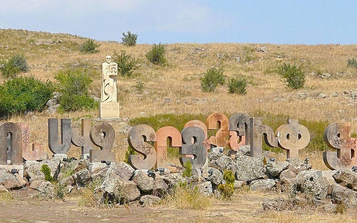 Görselde, açık bir alanda büyük taş harflerle yazılmış bir yazı ve arka planda bir heykel bulunuyor. Taş harfler, Ermenice alfabenin büyük harflerinden oluşuyor ve her biri farklı renkte ve dokuda. Harfler, doğal taşlardan yapılmış gibi görünüyor ve yere sabitlenmişler. Harflerin etrafında kaya parçaları ve küçük çalılar yer alıyor.  Arka planda, insan figürü şeklinde oyulmuş bir heykel var. Heykel, elinde bir kitap veya tablet tutan bir kişiyi tasvir ediyor. Bu alan, doğayla iç içe, açık ve geniş bir arazide bulunuyor. Arka plandaki hafif sararmış otlar, bölgenin kırsal ve doğal bir alan olduğunu gösteriyor. Heykel ve harfler, kültürel bir anıt ya da alfabe anıtı gibi bir yapıya işaret ediyor.