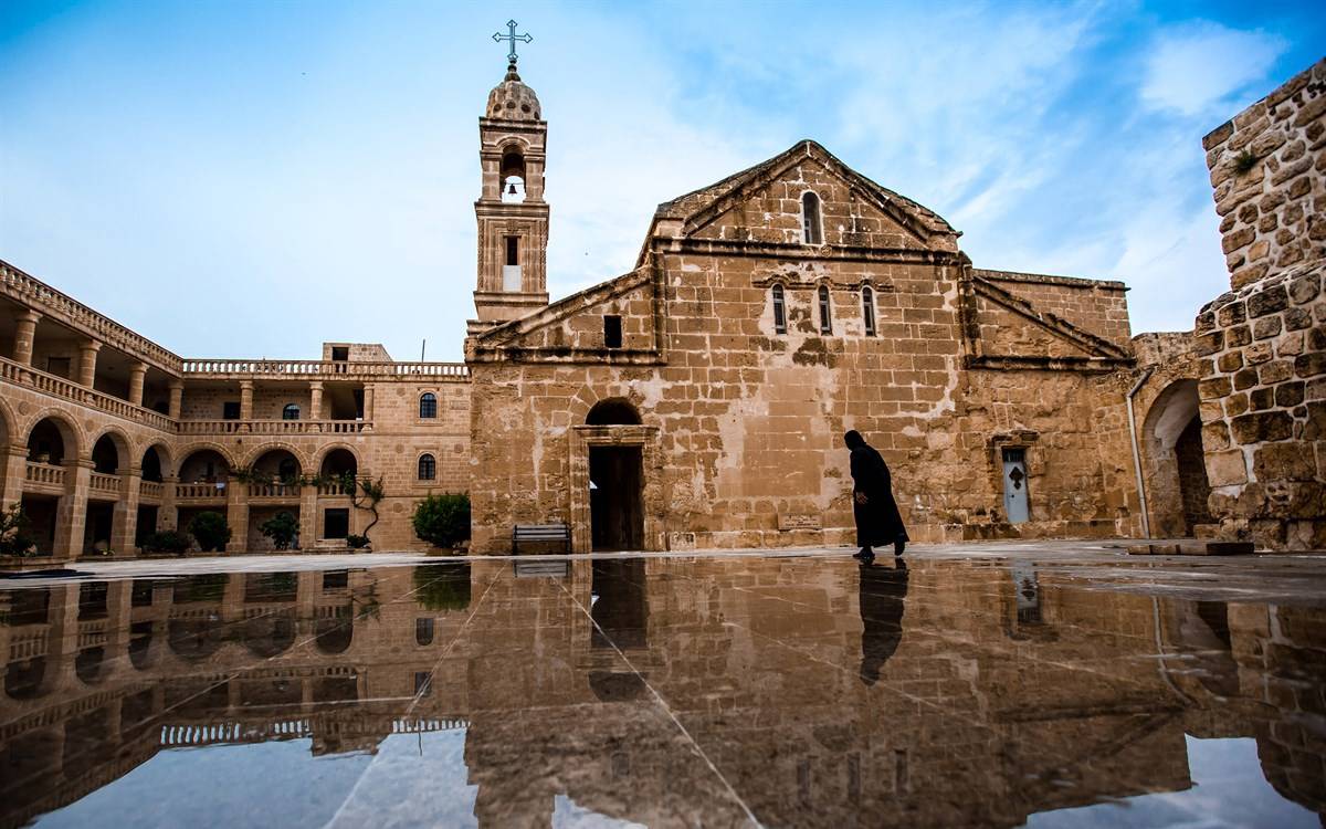 Tarihi Süryani Mor Yakup Manastırı. Manastır, taş duvarlarla inşa edilmiş olup, klasik mimari unsurları taşımaktadır. Sol tarafta kemerli geçitler ve üst katta balkonlar bulunmaktadır. Ortada ise büyük bir taş bina, kilise yapısı ve bir çan kulesi yer alıyor. Çan kulesinin üstünde büyük bir haç işareti dikkat çekiyor. Zeminde hafif su birikintisi var ve binanın yansıması suya düşmüş. Görselin sağ kısmında, siyah cübbe giymiş bir din adamı ya da keşiş, manastırın içinde yavaşça yürürken görüntülenmiş. Gökyüzü bulutlu ve hafif aydınlık.