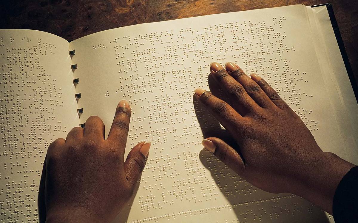 Bu görselde, Braille alfabesiyle yazılmış bir metni okuyan iki el görülüyor. Ellerin sahibi, parmak uçlarıyla sayfadaki kabartmalı noktaları hissederek okuma yapıyor. Sayfanın tamamında Braille alfabesiyle yazılmış metin bulunuyor ve eller dikkatlice bu noktaların üzerinde hareket ediyor. Arka planda ahşap bir yüzeyin küçük bir kısmı da fark ediliyor. Görsel, dokunarak okumanın önemini ve Braille alfabesinin kullanımını yansıtıyor.