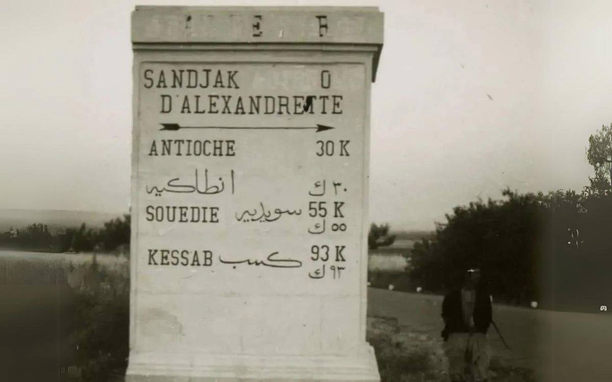 Görselde bir yol tabelası bulunuyor. Tabela dikdörtgen şeklinde ve üzerinde hem Latin hem de Arap harfleriyle yazılar var. Üst kısımda "Sandjak d'Alexandrete" yazıyor ve bu yazının yanında sağa doğru bir ok işareti bulunuyor. Hemen altında "Antioch" (Antakya) yazısı var ve karşısında 30 kilometre anlamına gelen "30 K" ifadesi bulunuyor. Bu bilgi Arapça harflerle de tekrar yazılmış. Tabelada ayrıca "Souedie" (Samandağ) 55 kilometre anlamında "55 K" olarak belirtilmiş. Yine bu bilgi de Arap harfleriyle yazılmış. Son olarak "Kessab" yazısı yer alıyor ve karşısında 93 kilometre anlamında "93 K" ifadesi var. Bu mesafe bilgisi de Arapça ile tekrarlanmış. Arka planda açık bir alan ve hafif ağaçlık bir manzara görünüyor. Sol tarafta, yol kenarında bir kişi silüeti yer alıyor, ancak detaylar net değil.