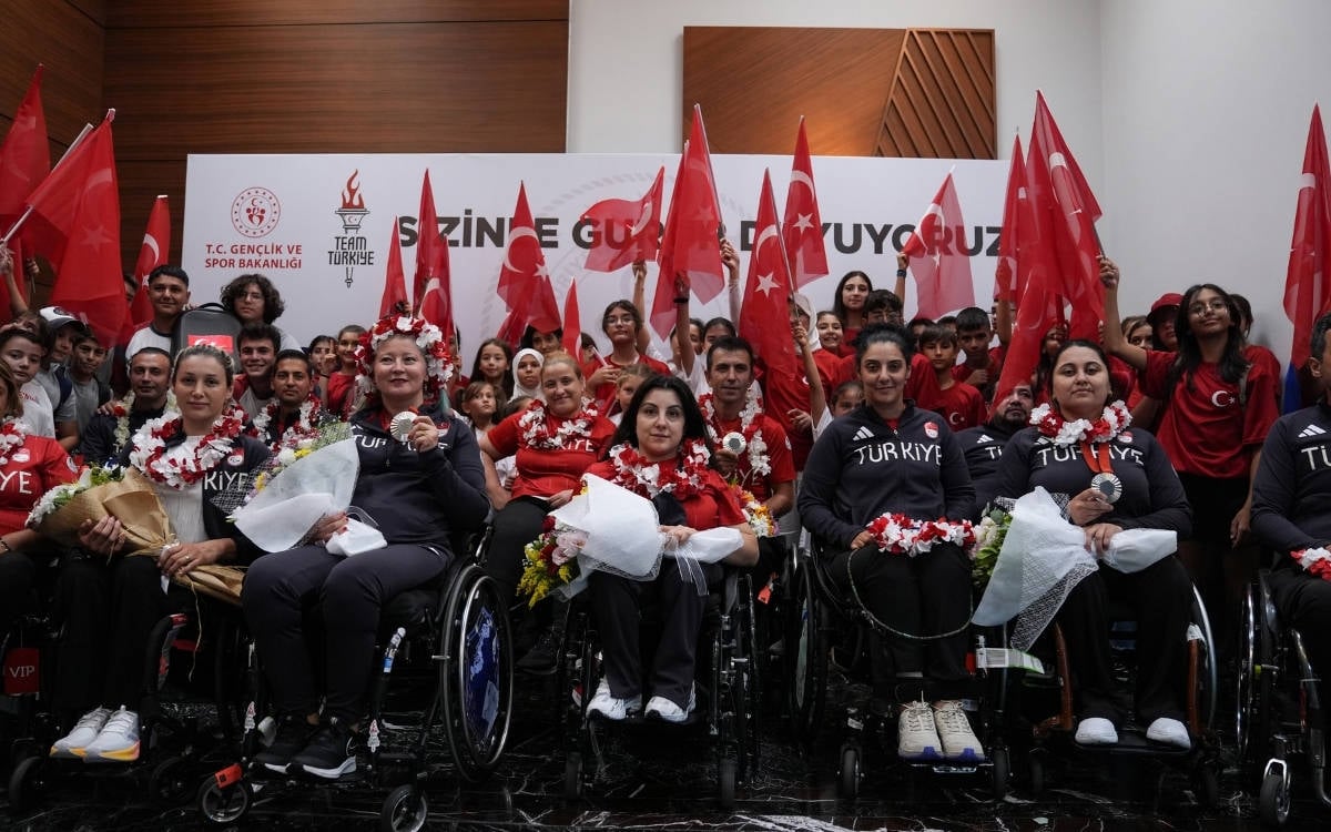 Görselde, Türkiye paralimpik milli takımı sporcuları ve taraftarlar bir arada görülmektedir. Ön sırada, tekerlekli sandalyede oturan kadın sporcular çiçeklerle ve madalyalarla poz vermektedir. Üzerlerinde "Türkiye" yazılı koyu renkli spor kıyafetleri bulunmaktadır. Sporcuların başlarında çiçekten taçlar yer almakta ve ellerinde başarılarını simgeleyen madalyalar veya ödüller bulunmaktadır.  Arka planda ise birçok kişi Türk bayraklarıyla onları desteklemektedir. Kırmızı tişörtler giymiş kalabalık, coşkulu bir şekilde bayraklarını sallamaktadır. Arkadaki duvarda "T.C. Gençlik ve Spor Bakanlığı" logosu, "Team Türkiye" yazısı ve "Sizinle Gurur Duyuyoruz" sloganı yazılıdır. Bu görsel, milli sporcuların başarısını kutlayan bir karşılama törenini veya bir etkinliği simgeliyor.